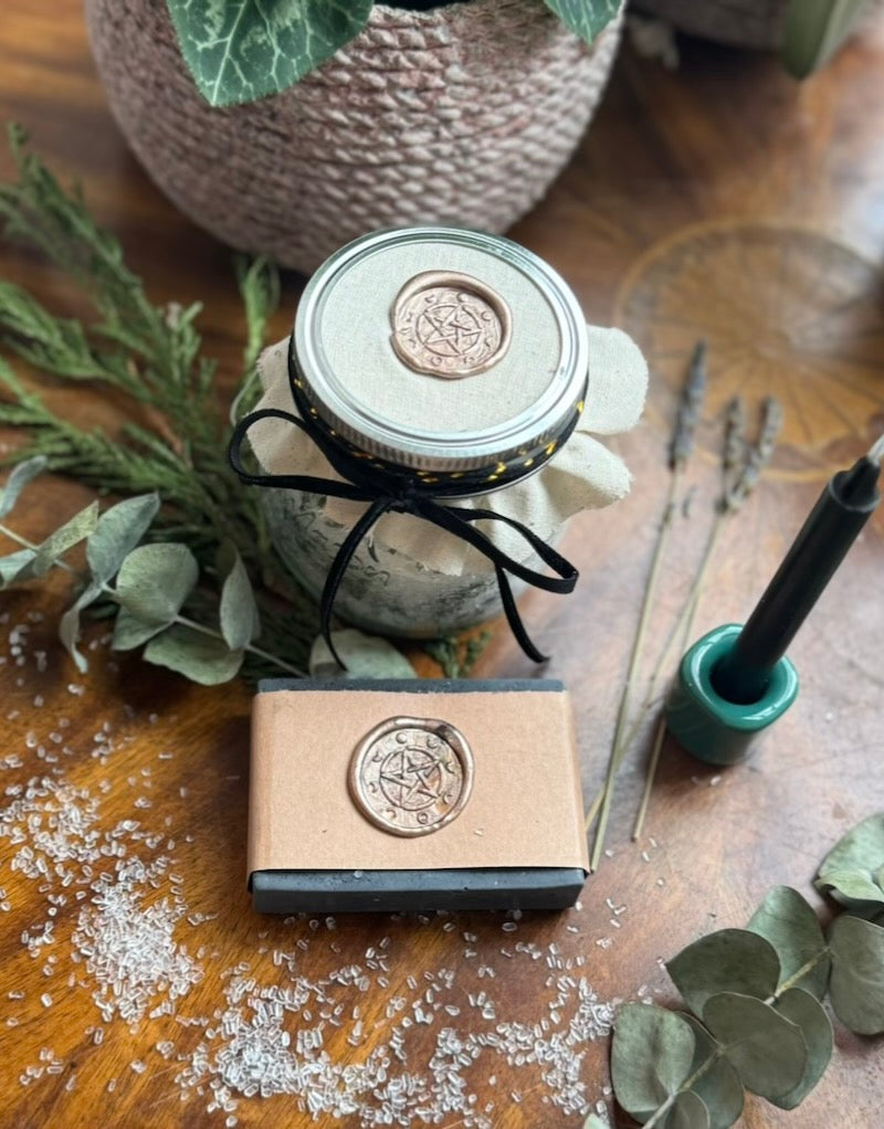 A Protection Ritual Bath Kit including a black candle, a small container of bath salts with herbs, and a bar of Charcoal Lavender soap, all arranged on a table with decorative leaves and a tool for lighting the candle.