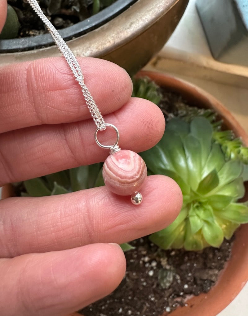 Hand holding a silver necklace with a pink bead in front of a potted plant